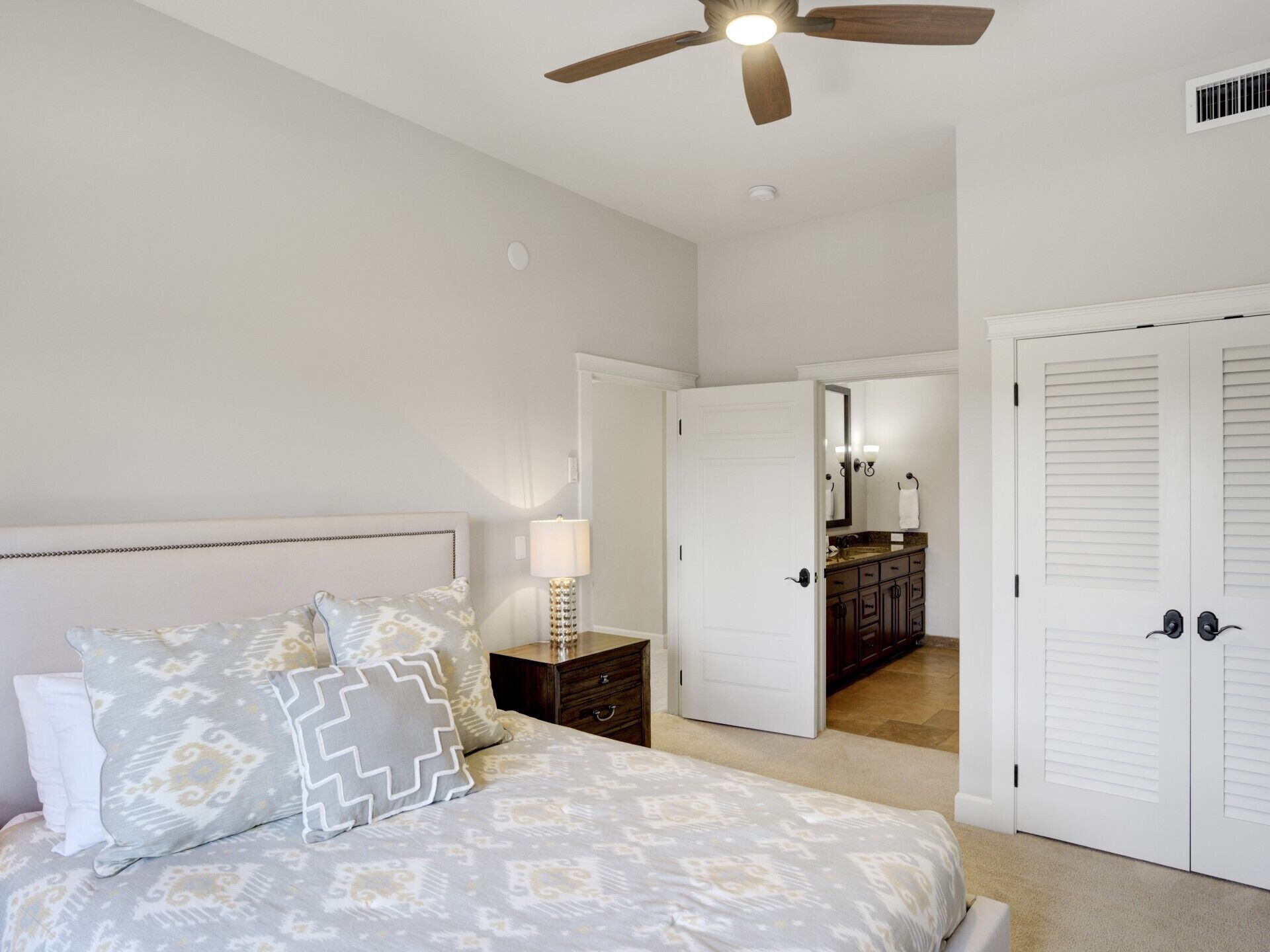 Un dormitorio con cama, mesita de noche, lámpara, ventilador de techo y vista al baño.