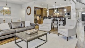 A spacious living room with a sofa, armchairs, and a coffee table. A kitchen with wooden cabinets and a dining area with bar stools is visible in the background. - Townhome, Groups Welcome - Seasons at Sandpoint (Sandpoint)