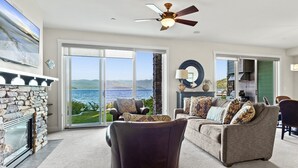 A spacious living room with a stone fireplace, a ceiling fan, and a large window overlooking a scenic view. - Townhome, Groups Welcome - Seasons at Sandpoint (Sandpoint)
