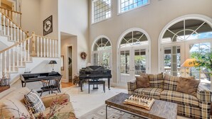 A spacious living room with a grand piano, a couch, and a coffee table with a chess set. - Broadmoor Estate Summer Rental - Designed for unforgettable family getaways. (Colorado Springs)