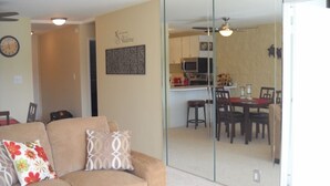A living room with a beige sofa, two patterned cushions, and a wooden coffee table. A glass partition separates the living room from a kitchen area with a dining table and chairs. - 195-295/night, Oceanfront with 2 Master Suites, Fully Stocked, Amazing View!!! (Wailuku)