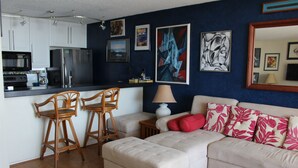 Ein Wohnzimmer mit einer Couch, einem Hocker und zwei Holzsesseln. Es gibt eine Küchenzeile mit Kühlschrank und Mikrowelle.