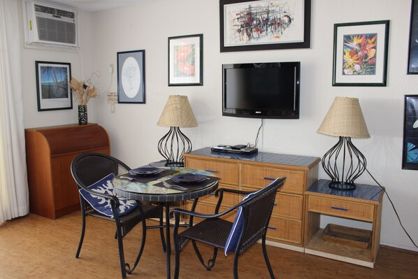 A compact living space with a dining table, chairs, a sofa, a TV, and artwork on the walls. - Kona Reef F13 - Nicely Renovated - Ocean View - Free WiFi, 3 minute walk to town (Kailua Kona)