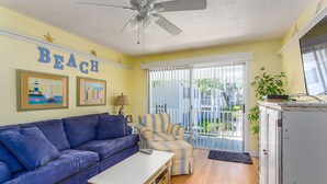 Ein Wohnzimmer mit einem blauen Sofa, einem gestreiften Sessel, einem weißen Couchtisch und einem Deckenventilator. An der Wand hängen zwei gerahmte Bilder, darüber das Wort "BEACH".