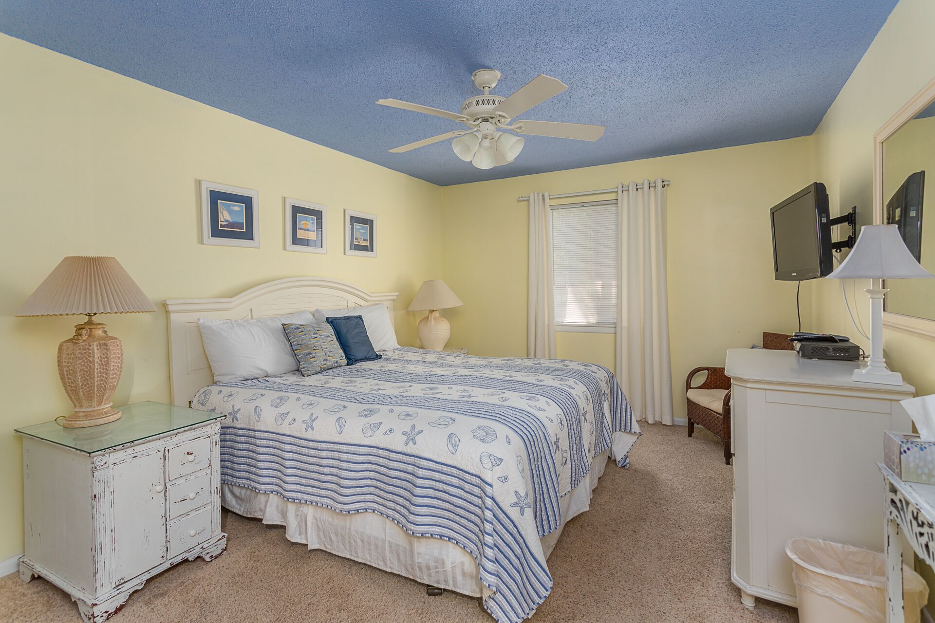 A bedroom with a bed, two bedside tables, a ceiling fan, a window with blinds, and a television on a stand.