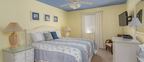 A bedroom with a bed, two bedside tables, a ceiling fan, a window with blinds, and a television on a stand.