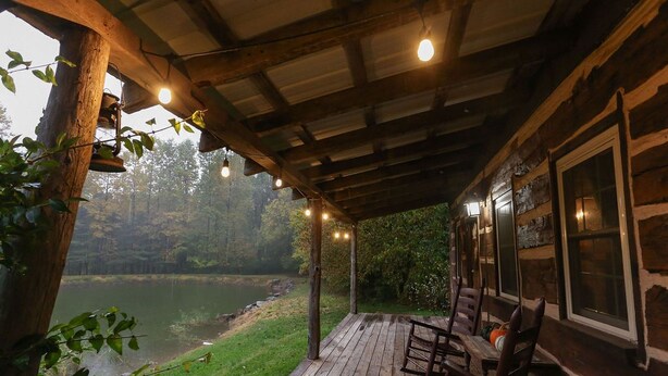 A wooden cabin with a deck overlooking a lake, featuring rustic wooden furniture and string lights.