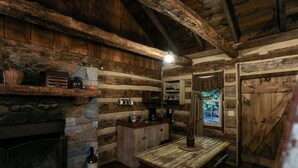 A rustic cabin interior with a stone fireplace, wooden shelves, a wooden table, and a window with a view of greenery.