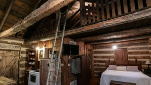 A rustic log cabin interior with a wooden staircase, a bed, a television, a stove, and a ladder.