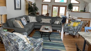 A spacious living room with a gray sectional sofa, a patterned armchair, and a coffee table. There are multiple windows providing natural light, and a ceiling fan is visible. - Spacious home,saltwater heated pool, game room, volleyball,ocean views & Tennis (Corolla)