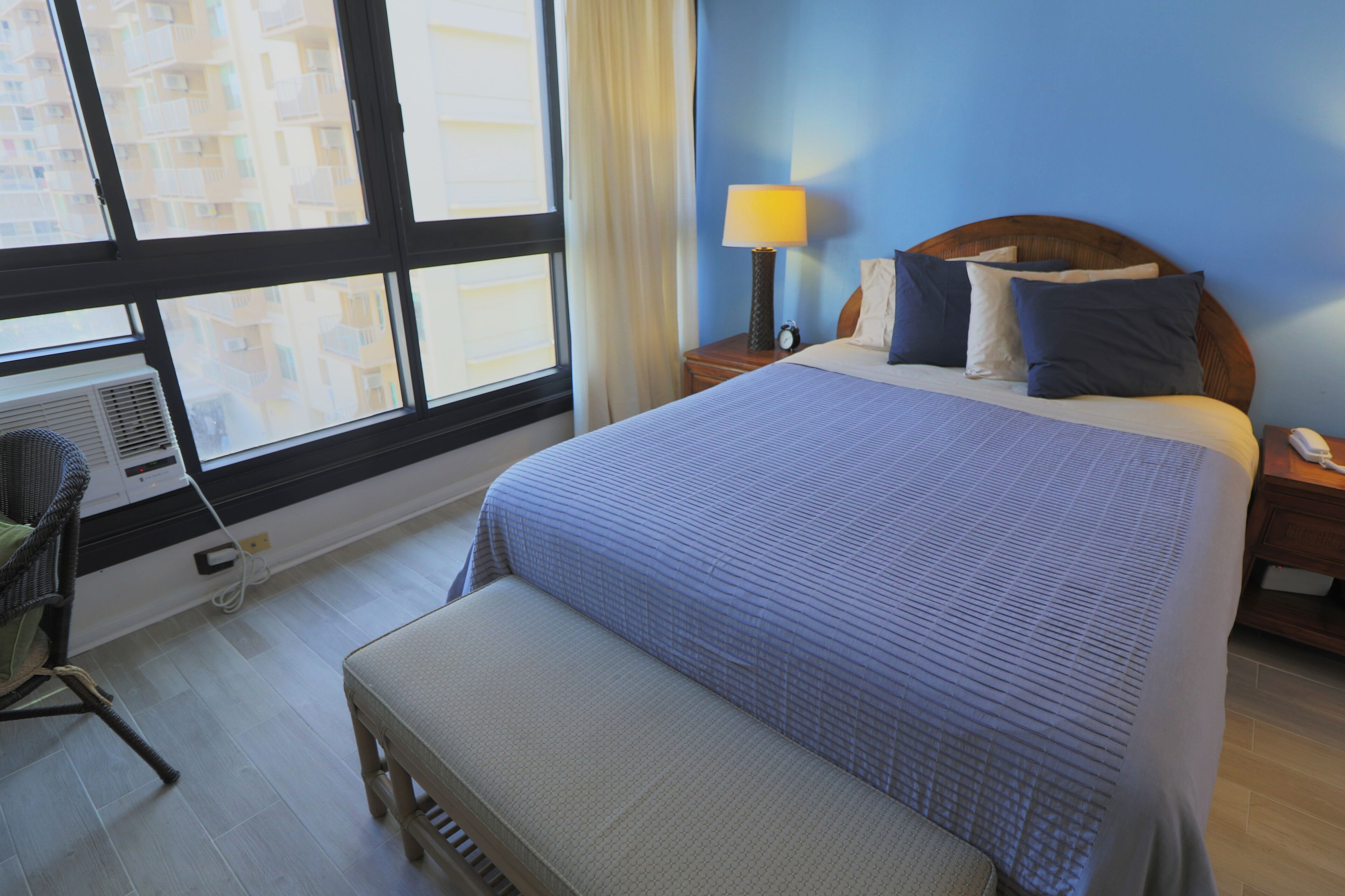 A hotel room with a large bed, a bedside table with a lamp, a chair, and a window with a view of buildings.