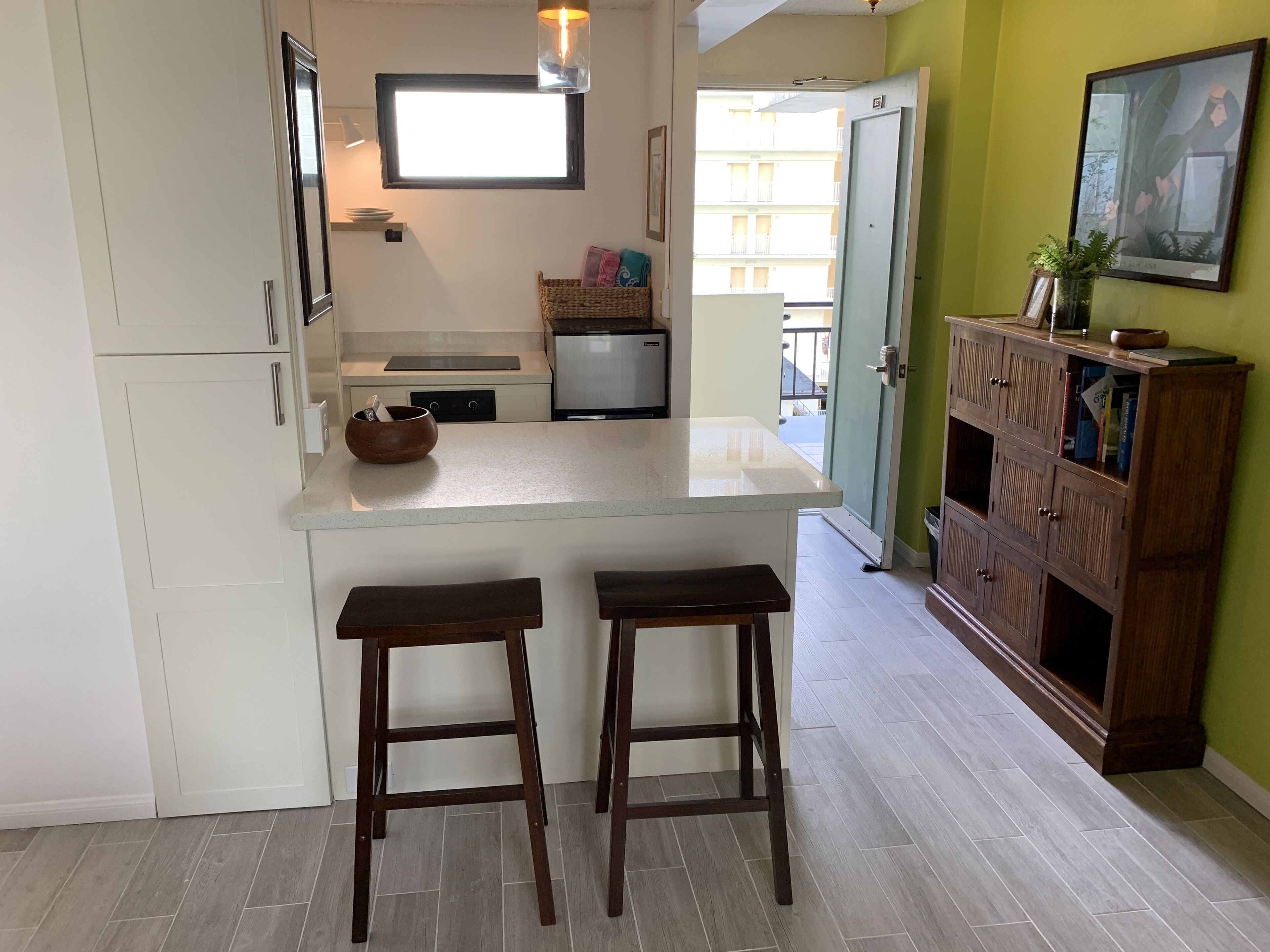 A kitchen with a central island, two stools, a refrigerator, a microwave, and a window.
