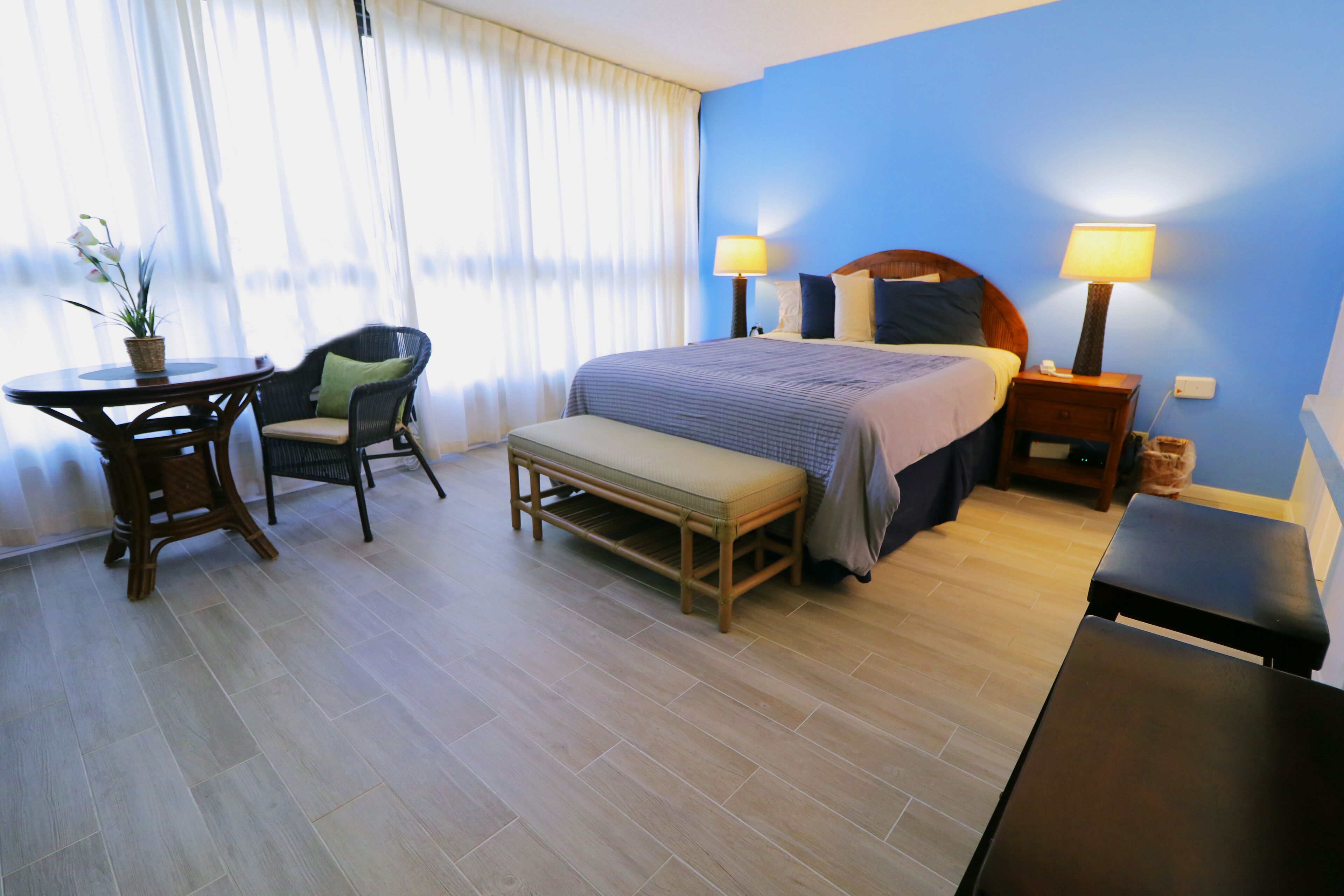 A hotel room with a bed, bedside tables, a bench, a chair, a table, and a potted plant.