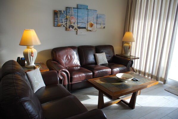 Een woonkamer met een bruine leren bank, twee bijzettafels met lampen, een salontafel en een wand met een schilderij in nautische stijl.
