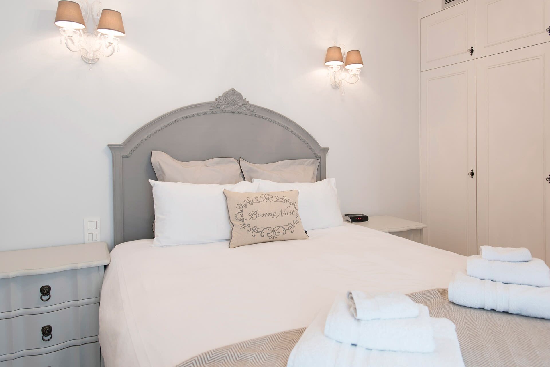 Una cama bien hecha con almohadas y un cojín decorativo, una mesita de noche con una lámpara, y un juego de toallas en el suelo.