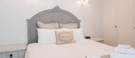 Una cama bien hecha con almohadas y un cojín decorativo, una mesita de noche con una lámpara, y un juego de toallas en el suelo.