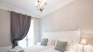 A hotel room with two beds, a chandelier, a window with curtains, and a bedside table with a lamp. - Stunning & Spacious in Superb Location with Balcony & Eiffel Views (Paris)