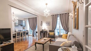 A spacious living room with a sofa, coffee table, and a TV. There is a balcony with a view, and a kitchen area is visible in the background. - Stunning & Spacious in Superb Location with Balcony & Eiffel Views (Paris)