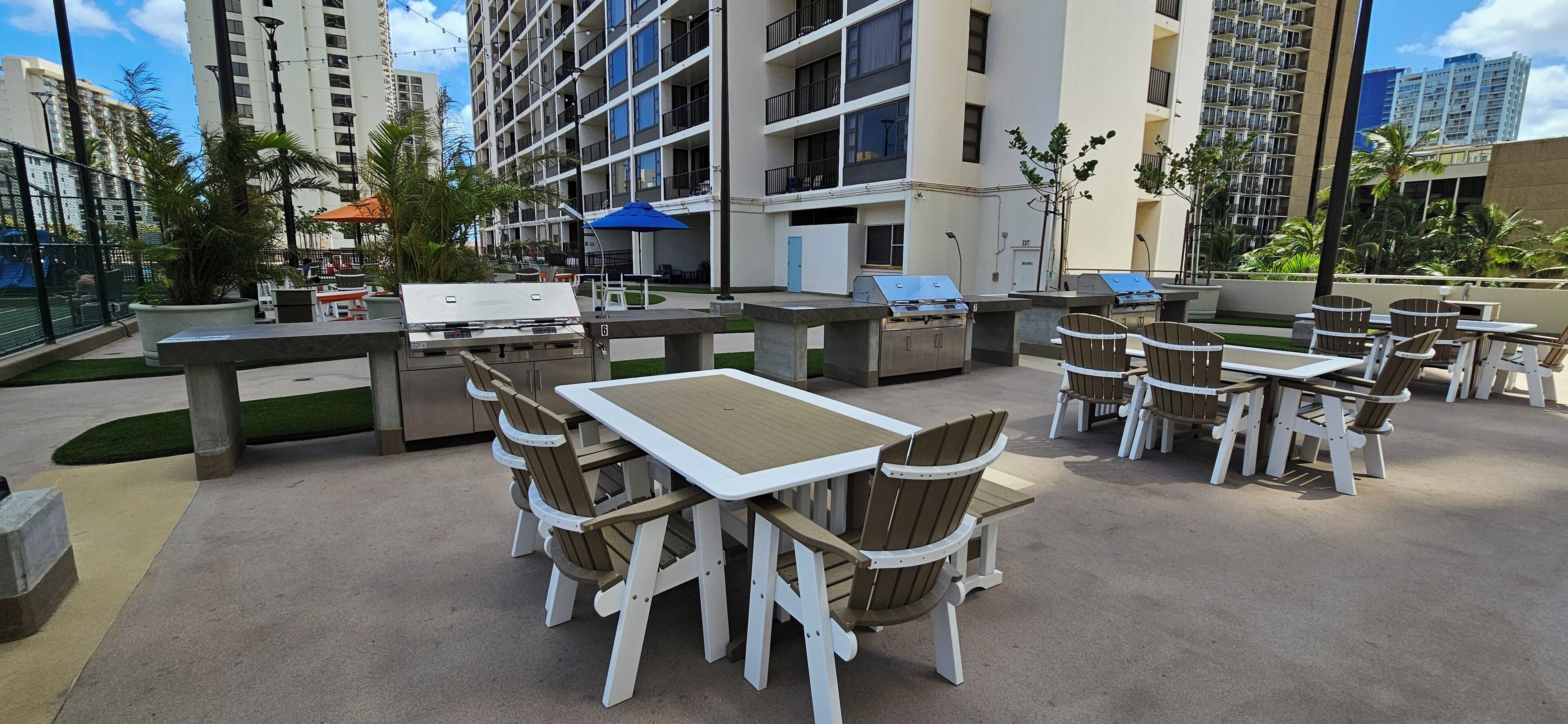 An outdoor patio area with tables and chairs, a barbecue grill, and a pool in the background.