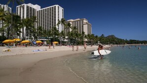 Una playa con palmeras, sombrillas y un hotel al fondo.