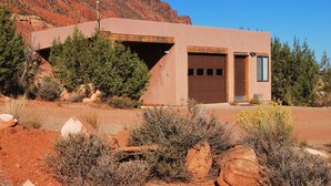 A modern two-story building with a garage, surrounded by desert vegetation and a mountain backdrop. - Cozy Adobe with Hot Tub and Fabulous Views in Outdoor Paradise. (Castle Valley)