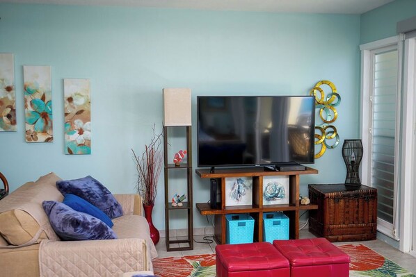 A living room with a flat-screen TV, a sofa, a red ottoman, and artwork on the walls. - Ocean and Surfer Beachfront/AC/Elevator (Kailua-Kona)