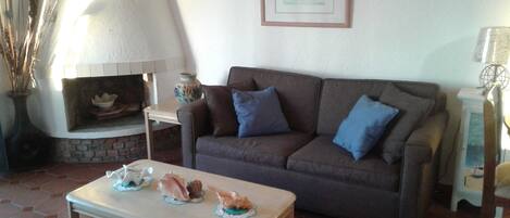 A living room with a brown sofa, two blue cushions, a small table with decorative shells, a fireplace, and a framed picture on the wall.
