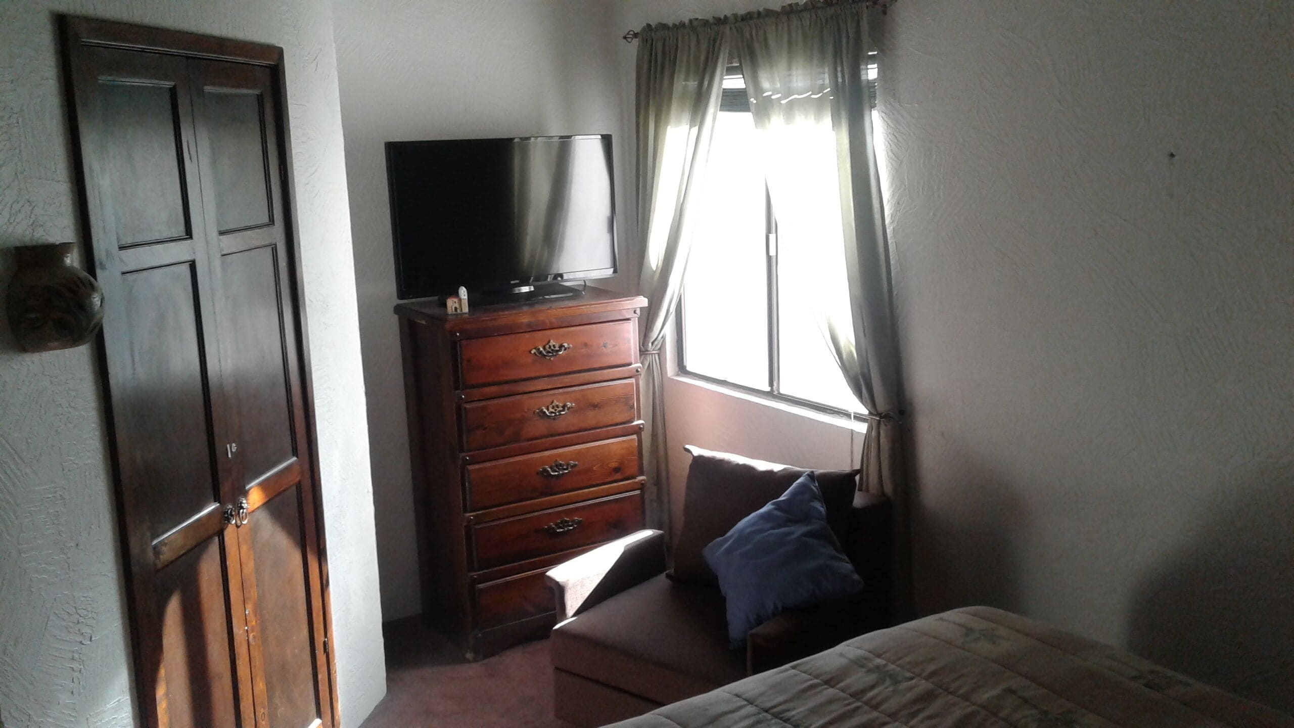A bedroom with a wooden dresser, a television on top, a bed with a blue pillow, and a window with curtains.