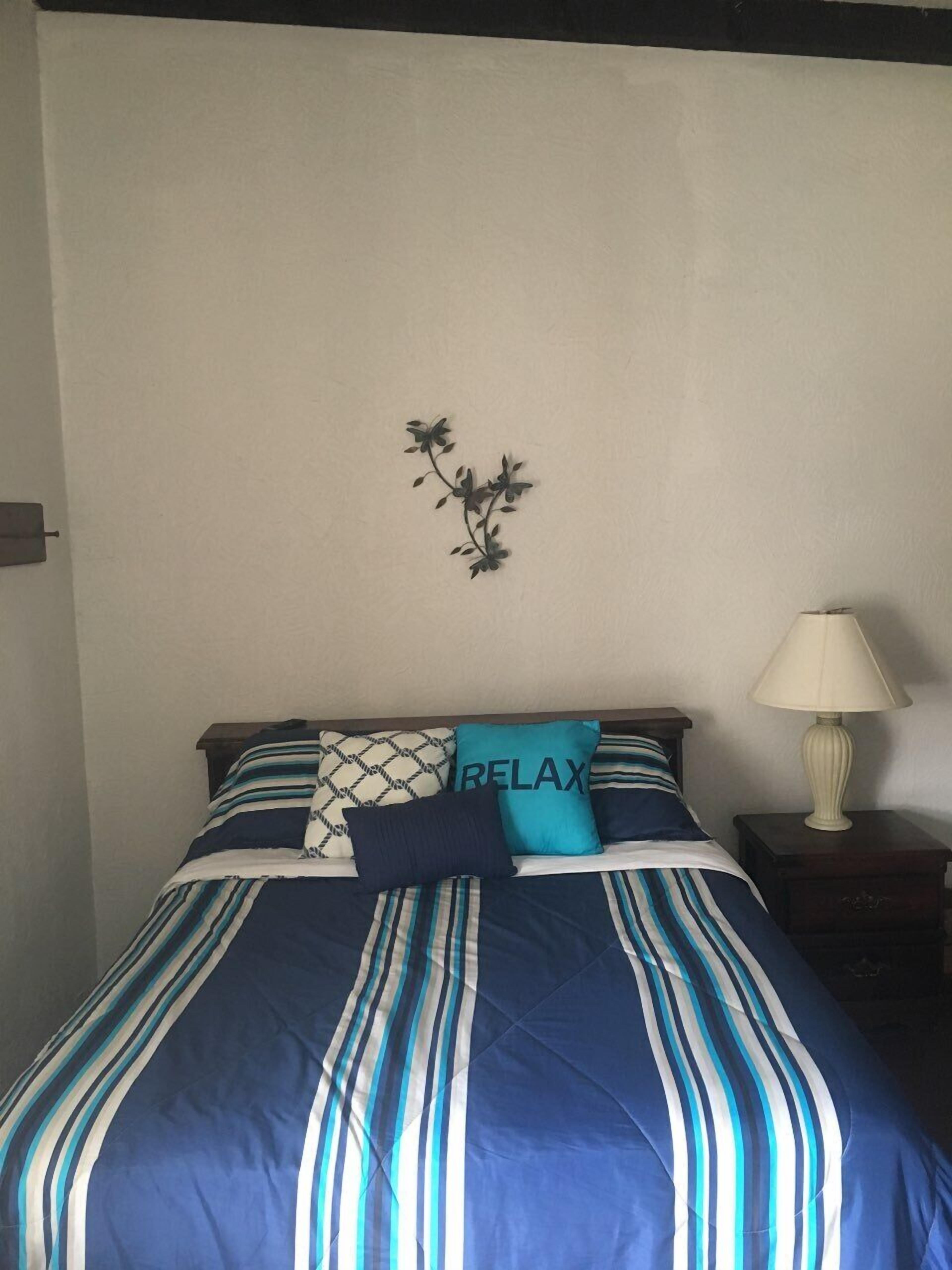 A bed with striped bedding, a pillow with the word "RELAX", a wall-mounted plant, and a bedside lamp.