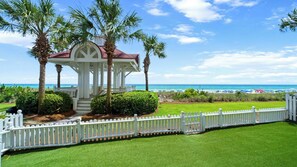 A gazebo with a red roof, surrounded by palm trees and a white picket fence, overlooking a beach with umbrellas. - Surfin' Sea | 1 PM Check In! | Modern Updated Gulf-Front Home | Private Pool | FREE Beach Chairs Within Steps | 6-Seat Golf Cart in Garage! | Stunning Views! (Miramar Beach)