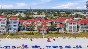A coastal residential area with beachfront properties featuring balconies, red roofs, and a sandy beach with umbrellas. - Surfin' Sea | 1 PM Check In! | Modern Updated Gulf-Front Home | Private Pool | FREE Beach Chairs Within Steps | 6-Seat Golf Cart Included! | Stunning Views! (Miramar Beach)