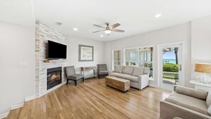 A spacious living room with a fireplace, a ceiling fan, a large TV, and a view of the ocean through the glass doors. - Surfin' Sea | 1 PM Check In! | Modern Updated Gulf-Front Home | Private Pool | FREE Beach Chairs Within Steps | 6-Seat Golf Cart in Garage! | Stunning Views! (Miramar Beach)