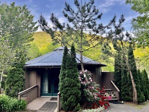 A cabin with a metal roof, surrounded by evergreen trees and flowering shrubs.