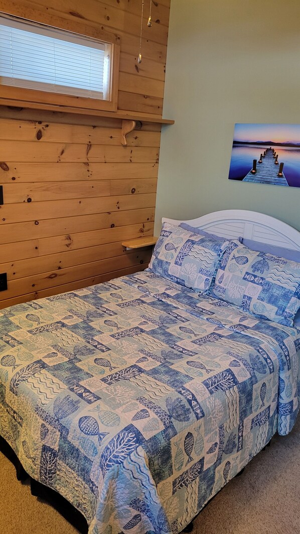 A bedroom with a wooden headboard, a bed with a blue and white patterned bedspread, a wall-mounted shelf, a window with blinds, and a painting of a pier.