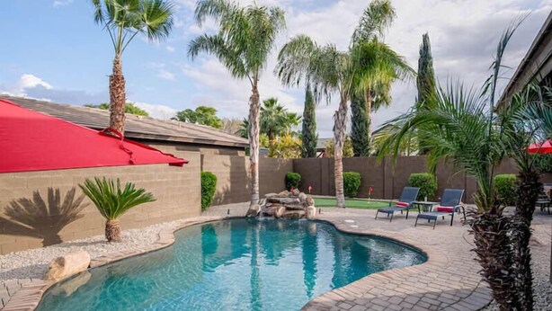 A swimming pool with a brick border, palm trees, and a red canopy.