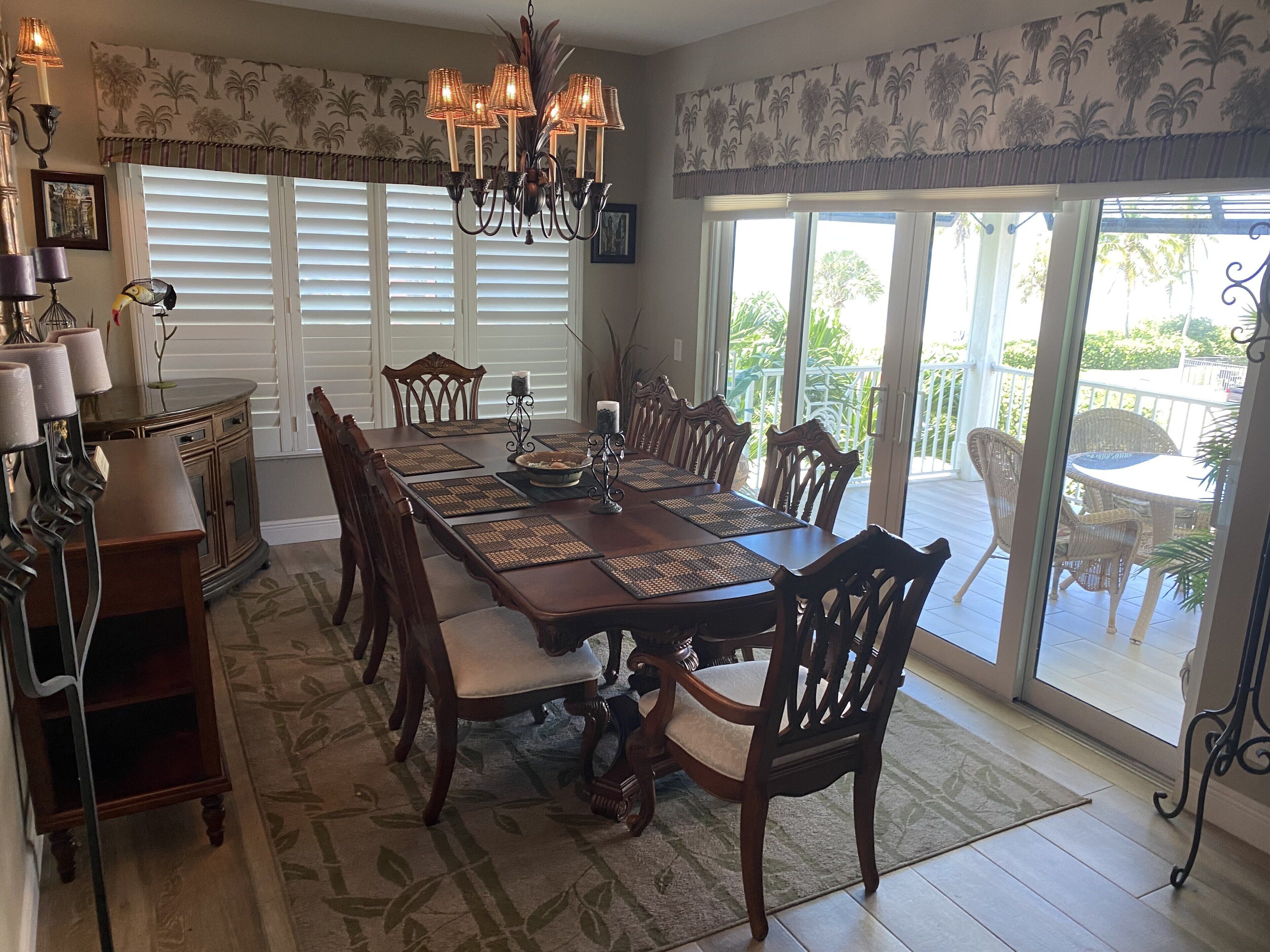 A dining area with a long table, chairs, and a chandelier. There is a window with blinds and a view of the outdoors.