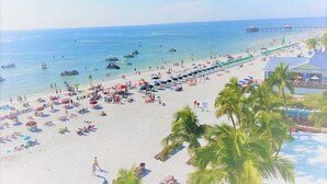 Una playa con filas de tumbonas y sombrillas, un muelle que se adentra en el agua y barcos.