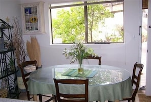 Une table de salle à manger ronde, avec un plateau en verre et une nappe verte, entourée de chaises en bois. Une fenêtre donne sur un paysage verdoyant.