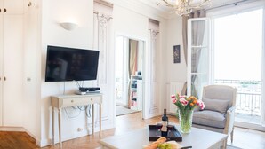 A living room with a flat-screen TV, a dining table with a bottle of wine, glasses, and a loaf of bread. There is a chair and a vase with flowers on the table. - Dream-Come-True With Breathtaking Eiffel & Paris Views + Terrace (Paris)