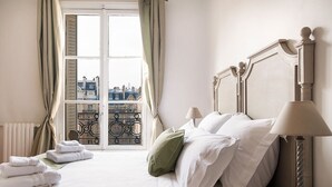 A hotel room with a bed, two bedside lamps, a window with curtains, and a view of buildings. - Dream-Come-True With Breathtaking Eiffel & Paris Views + Terrace (Paris)