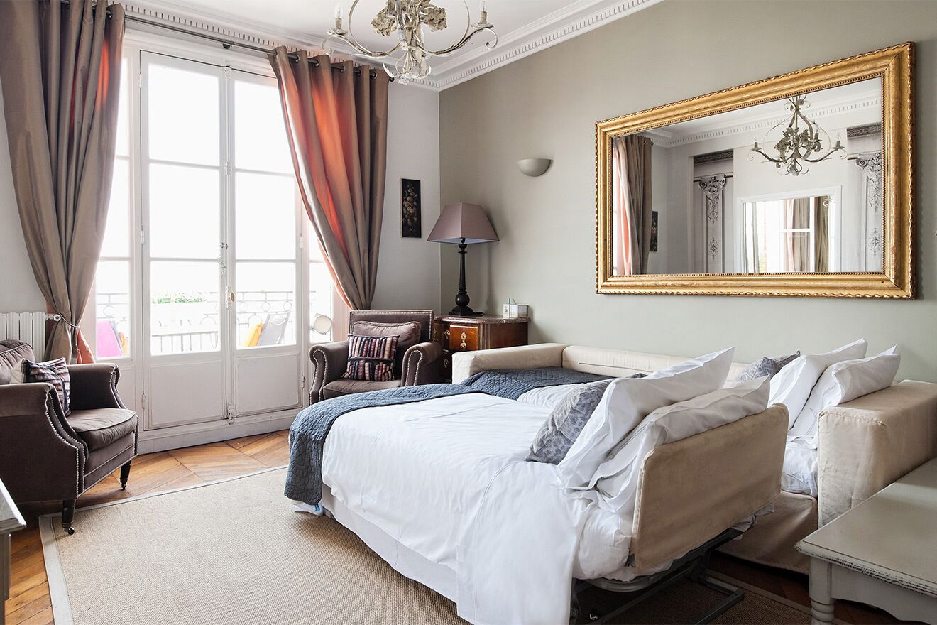 A bedroom with a large bed, a sitting area with a chair and a table, a large mirror, and a window with curtains.