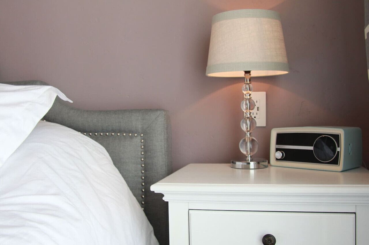 Un lit bien fait, avec une tête de lit grise, une table de chevet blanche sur laquelle se trouve une radio, et une lampe dotée d’un abat-jour à rayures.