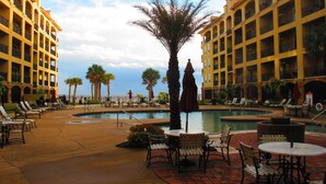 Área de piscina en un resort, con sillones y mesas, rodeada de palmeras y un edificio con balcones.