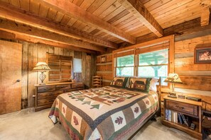 A cozy log cabin bedroom with a large bed, wooden dresser, and a window with a view of greenery.