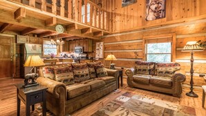 A cozy log cabin interior with a wooden dining table, two sofas with animal-themed cushions, a kitchen area, and a large window. - Fireplace, firepit, hot tub, minutes from Ski Resort - Low rates, no inflation ! (Gatlinburg)