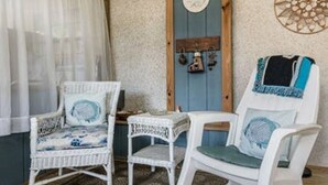 Une pièce comprenant trois chaises en osier blanches, dont une avec un coussin bleu et une autre avec un coussin vert. On y trouve une porte en bois ornée d’un miroir rond et une petite étagère sur laquelle sont disposés des objets décoratifs.