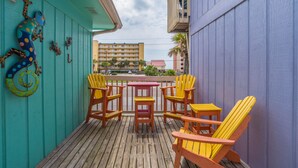 A wooden deck with yellow Adirondack chairs and a small table, a teal wall with a lizard decoration, and a view of buildings in the background. - 2 Bedroom, Across the street from the beach! & Pup-Friendly! (Gulf Shores)