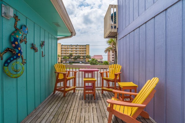 A wooden deck with yellow Adirondack chairs and a small table, a teal wall with a lizard decoration, and a view of buildings in the background. - 2 Bedroom, Across the street from the beach! & Pup-Friendly! (Gulf Shores)