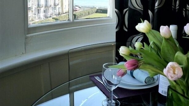 A dining area with a table set for two, a window with a view of buildings and the sea, and a vase with tulips. - Romantic with Sea View Sleeps 2-4 (Brighton)