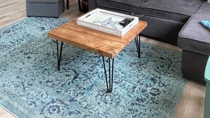 Ein Wohnzimmer mit grauem Sofa, einem Holztisch, einem blauen Teppich mit Muster, einem grauen Sessel, einem Beistelltisch mit Lampe und einer Küche im Hintergrund.